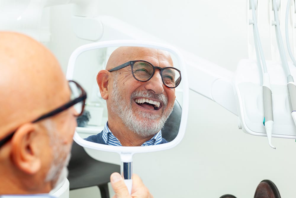 Man sitting in dentist chair looking into mirror and smiling at his new dental implants